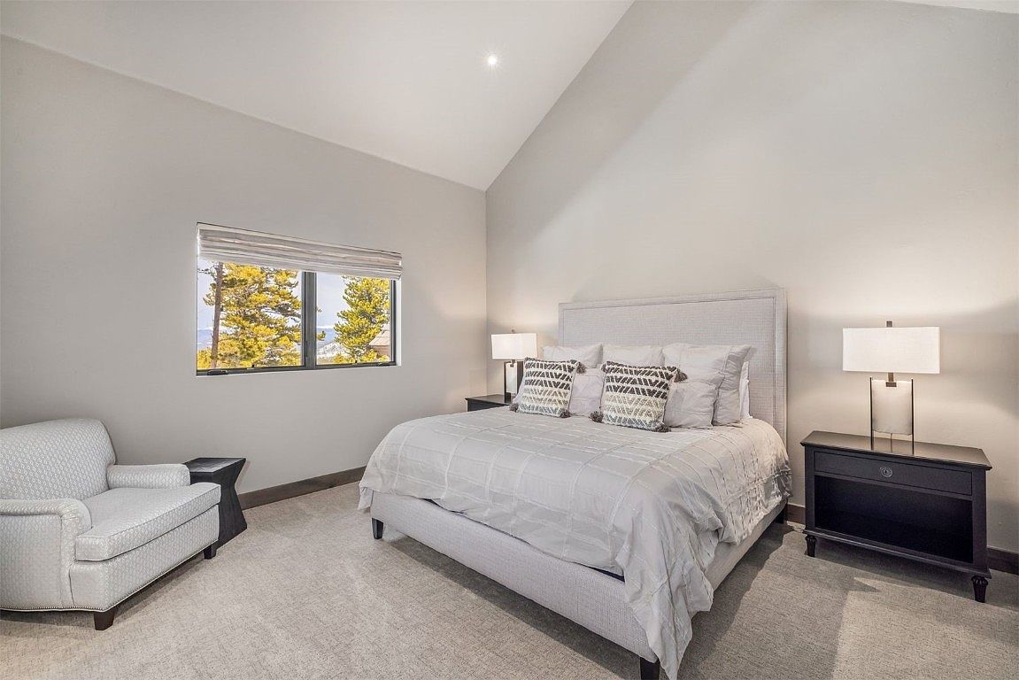 This is a well-lit guest bedroom featuring a queen-sized bed with a light-colored headboard and decorative pillows. A window provides natural light and a view of trees, while a bedside table with a lamp and an armchair offer additional comfort. The room has a neutral color palette and a cozy, inviting atmosphere.