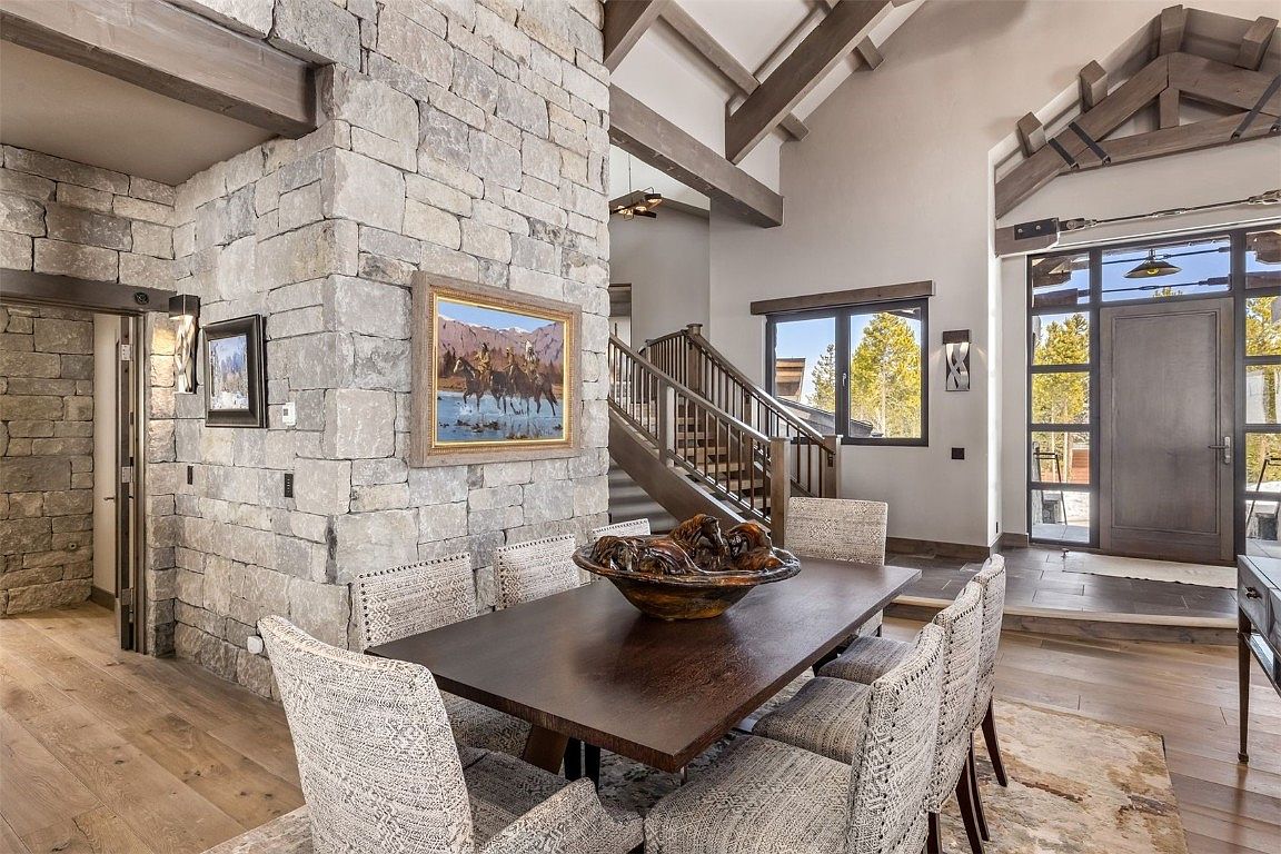 This interior shot showcases a dining room with a rustic yet elegant design. A large wooden dining table is surrounded by upholstered chairs, set upon a patterned rug. The room features a stone accent wall with artwork, wooden beams on the ceiling, and a staircase leading to the upper level, creating a warm and inviting atmosphere.