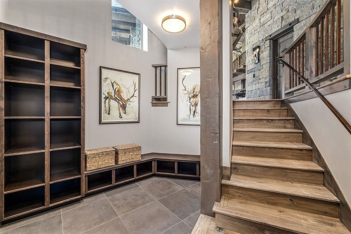 This interior shot showcases a hallway with wooden stairs leading upwards, complemented by a stone accent wall. A built-in bookshelf and bench seating provide functional storage and seating options. The space is illuminated by a modern ceiling light, and artwork adorns the walls, creating a warm and inviting atmosphere.
