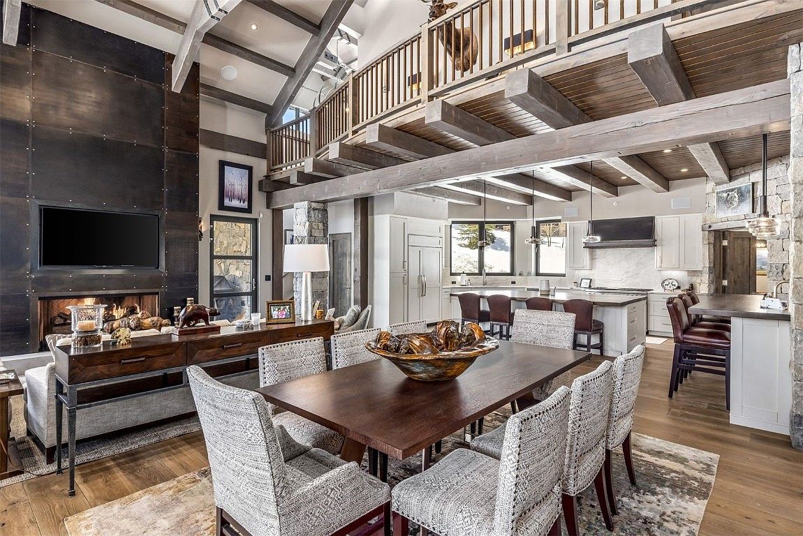 This interior shot showcases a dining area with a large wooden table surrounded by upholstered chairs, set on a patterned rug. The room features exposed wooden beams, stone accents, and a view into an open-concept kitchen. A striking metal fireplace surround with a mounted television adds a modern touch to the rustic-chic design.