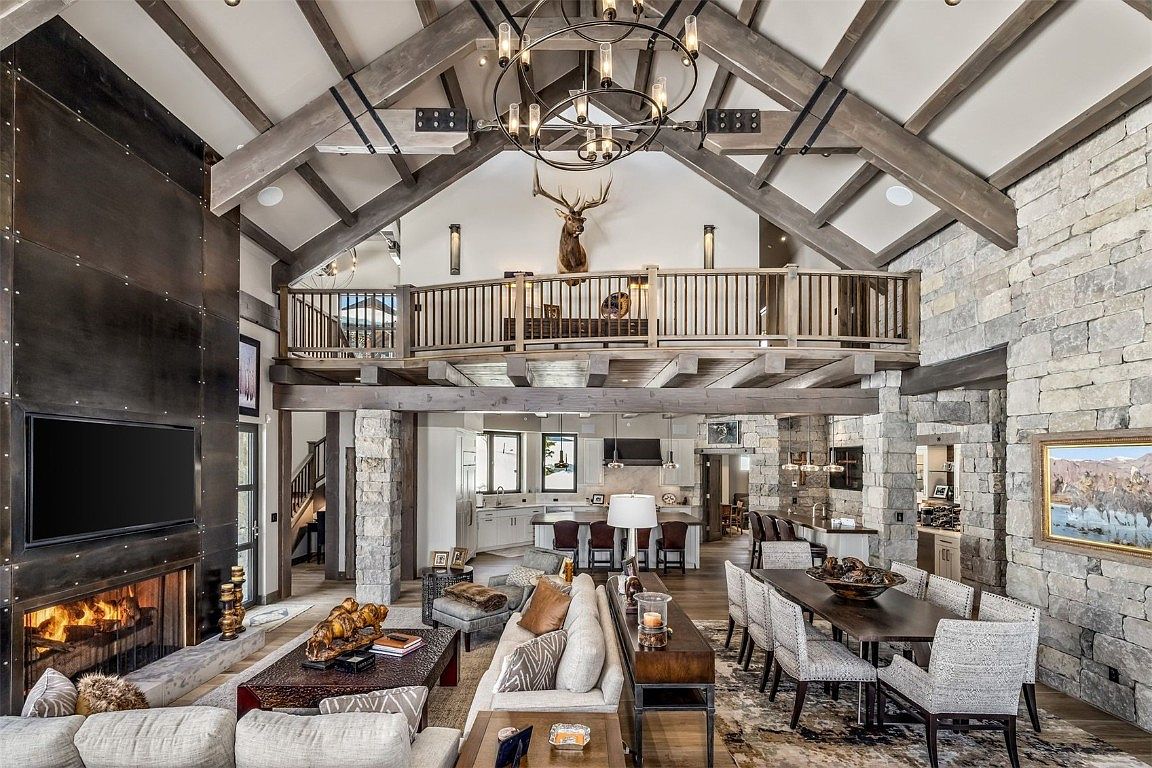 This is a spacious living room featuring a high vaulted ceiling with exposed wooden beams and a stone accent wall. A large fireplace with a dark metal surround is the focal point, and the room is furnished with comfortable sofas and a dining table. A second-floor balcony overlooks the living area, adding to the grandeur of the space.