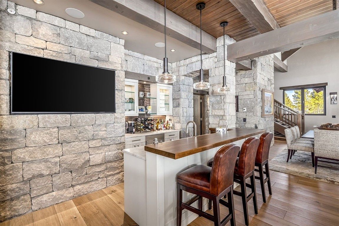 This interior shot showcases a stylish living room area featuring a stone accent wall with a large flat-screen TV. A bar area with wooden countertop and leather bar stools adds a touch of sophistication, complemented by modern pendant lighting. The hardwood floors and exposed wooden beams contribute to a rustic yet elegant ambiance.