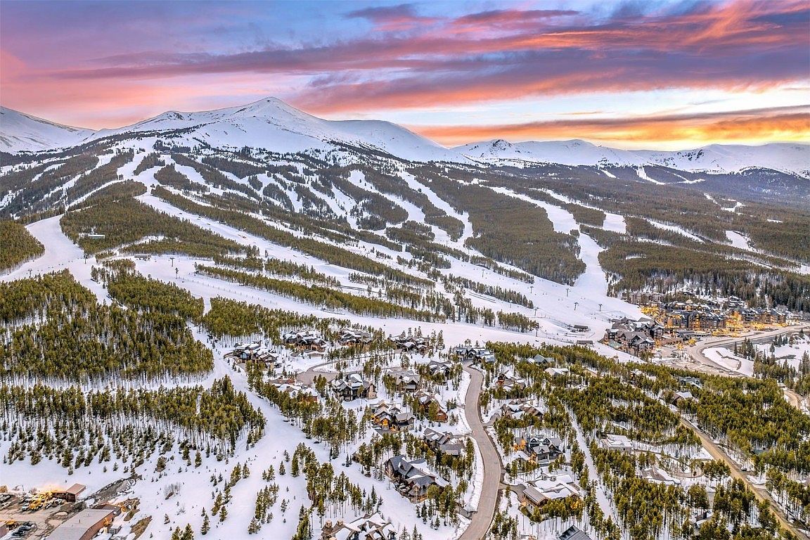 This aerial view showcases a stunning mountain resort community at sunset. The scene features snow-covered mountains with ski runs, a dense forest of evergreen trees, and luxury homes nestled among the trees. The sky is painted with vibrant hues of pink, orange, and purple, creating a picturesque and inviting atmosphere.