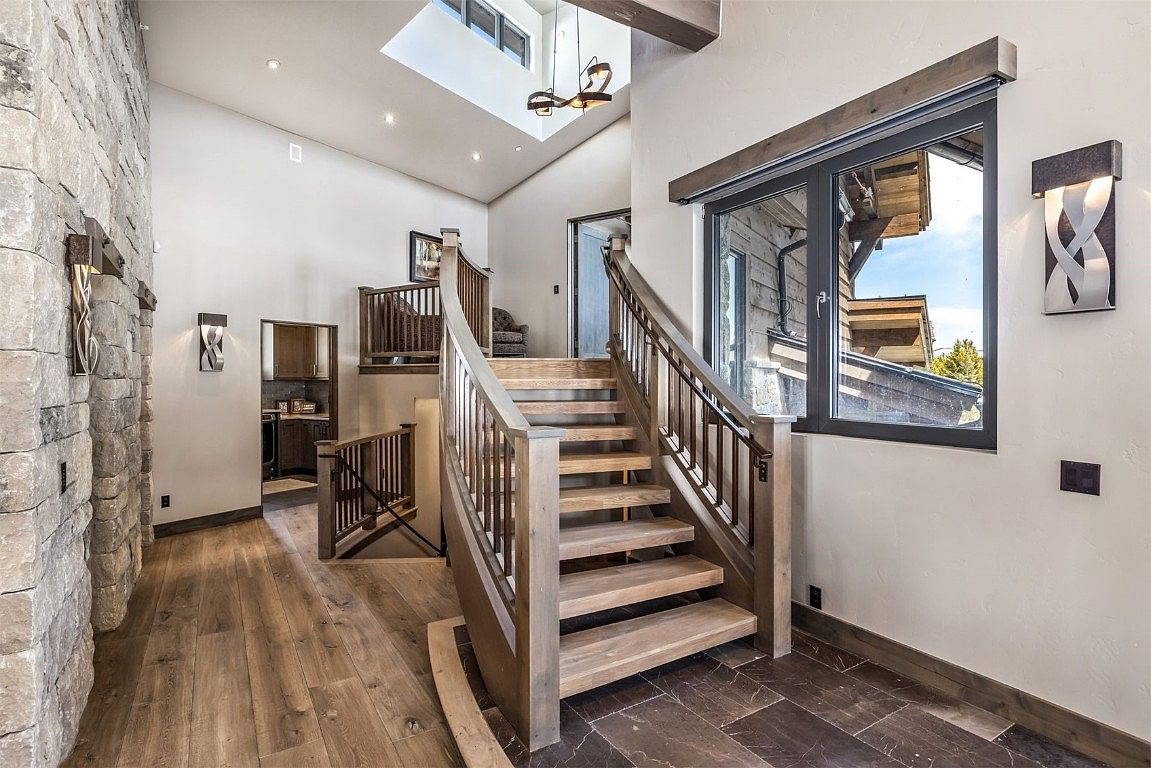 This interior shot showcases a grand hallway with a striking staircase. The staircase features light wood steps and dark wood railings, leading to an upper level. The hallway is adorned with a stone accent wall, modern light fixtures, and a mix of hardwood and tile flooring, creating a luxurious and inviting atmosphere.