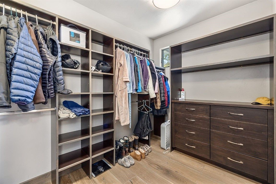 This is a well-organized walk-in closet featuring custom shelving and drawers. The closet contains a variety of clothing items, shoes, and accessories, all neatly arranged. The dark wood finish of the shelving and drawers adds a touch of elegance, while the light wood flooring provides a warm contrast.