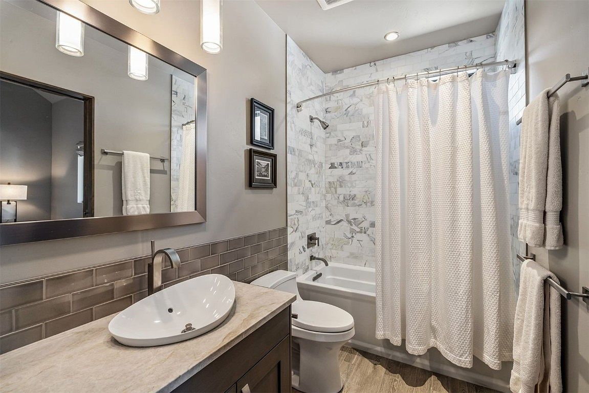 This is a well-lit bathroom featuring a modern design. The bathroom includes a vessel sink on a light-colored countertop, a toilet, and a bathtub with a white shower curtain. The walls are tiled with a combination of gray subway tiles and marble-patterned tiles, creating a clean and stylish look.