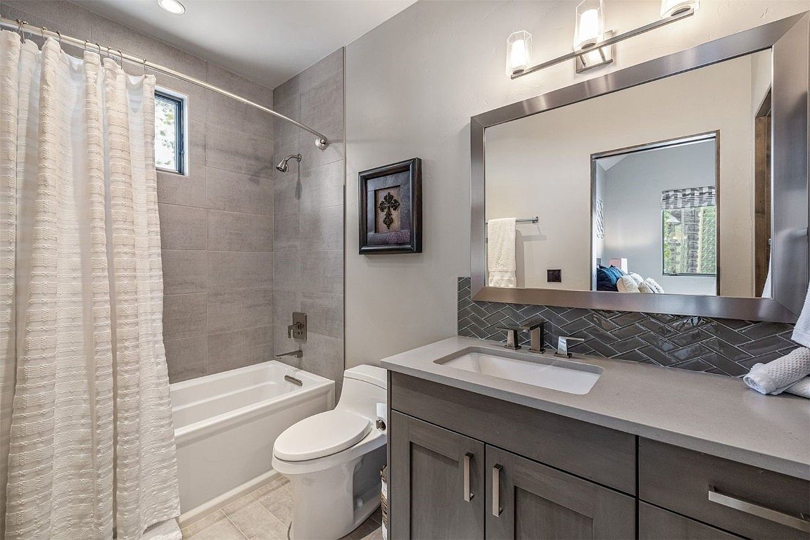 This is a well-lit guest bathroom featuring a bathtub with a shower curtain, a toilet, and a vanity with a sink. The walls are tiled in a neutral gray, and a framed artwork hangs above the toilet. A large mirror reflects the room, creating a sense of spaciousness.