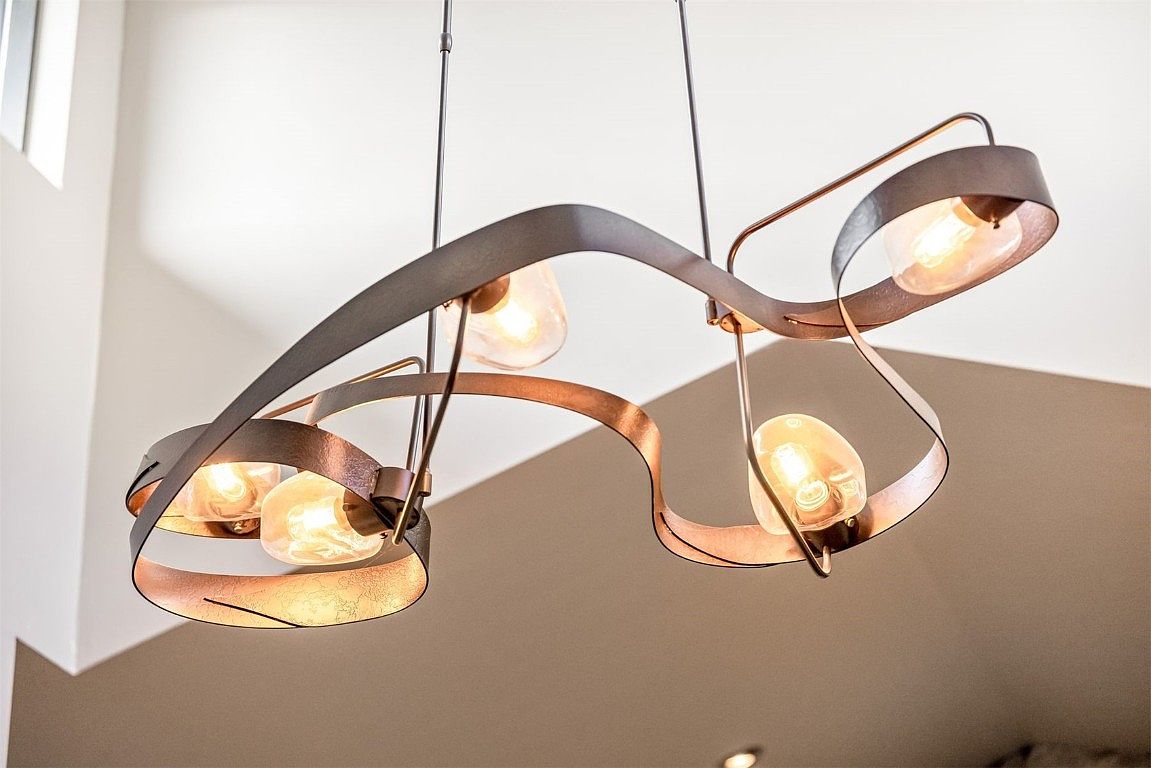 The image showcases a modern, artistic chandelier with a unique design featuring curved metal bands in a bronze or copper finish. The light fixture has multiple exposed bulbs, creating a warm and inviting glow. The overall impression is stylish and contemporary, adding a touch of sophistication to the space.