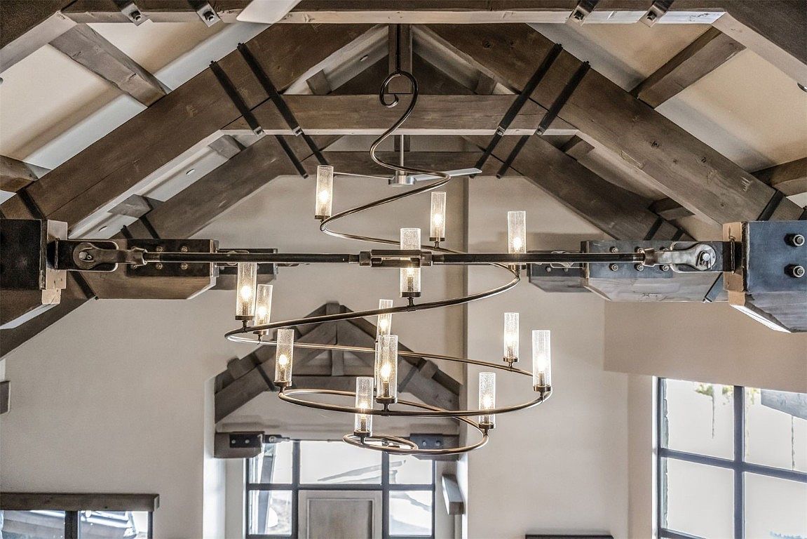 The image showcases a unique and modern chandelier suspended from a rustic, exposed beam ceiling. The chandelier features a spiral design with cylindrical glass lights, creating an elegant contrast against the industrial-style beams and metal hardware. The perspective is from below, emphasizing the height and architectural details of the space.