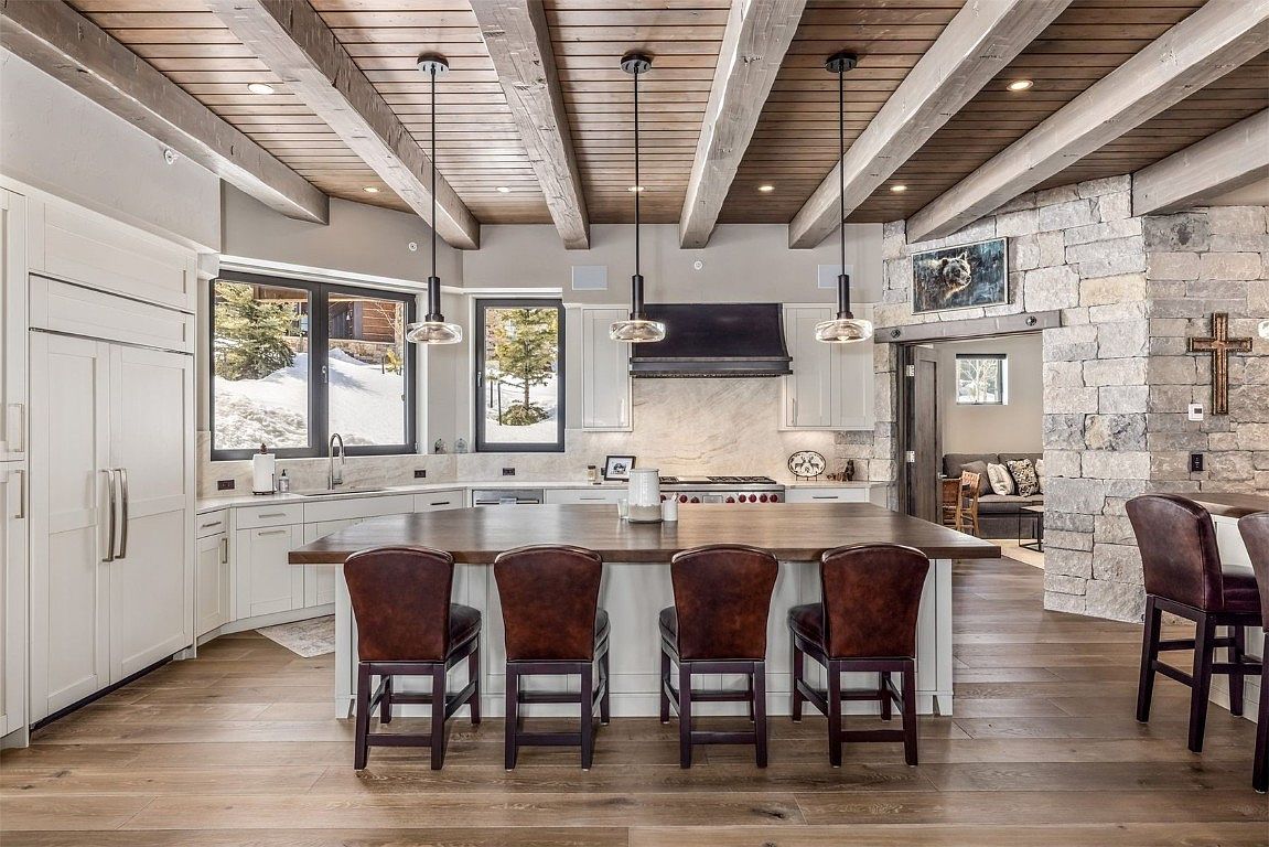 This is a well-lit kitchen featuring a large island with seating, white cabinetry, and stainless steel appliances. The ceiling has exposed wooden beams and pendant lighting. The kitchen opens to a living area with stone accents, creating a warm and inviting atmosphere.