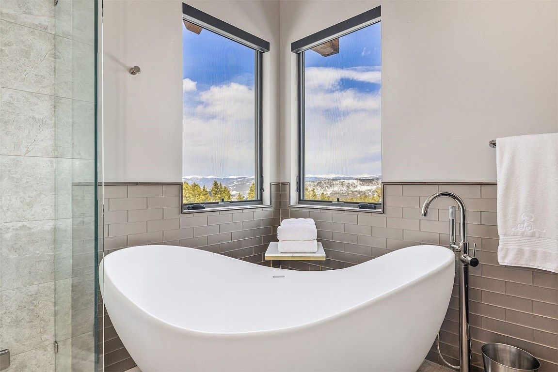 This is a luxurious primary bathroom featuring a freestanding white bathtub positioned beneath two corner windows that offer a scenic view of snow-capped mountains and trees. The walls are adorned with gray subway tiles, complemented by a modern chrome faucet and a neatly hung white towel. A glass-enclosed shower is visible to the left, adding to the bathroom's spa-like ambiance.