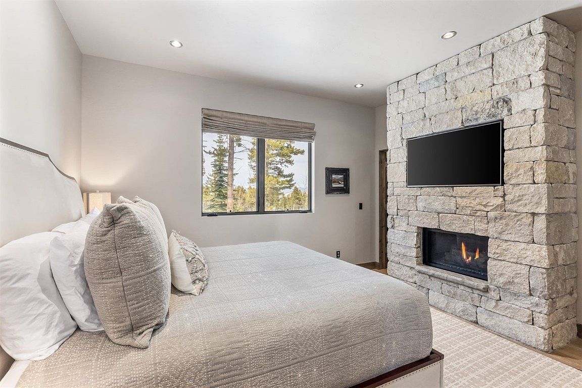 This is a primary bedroom featuring a large bed with neutral-toned bedding and pillows, a stone fireplace with a mounted television above, and a window with natural light. The room has a cozy and luxurious feel, with a focus on comfort and relaxation. The stone fireplace adds a rustic touch, while the overall design is modern and sophisticated.