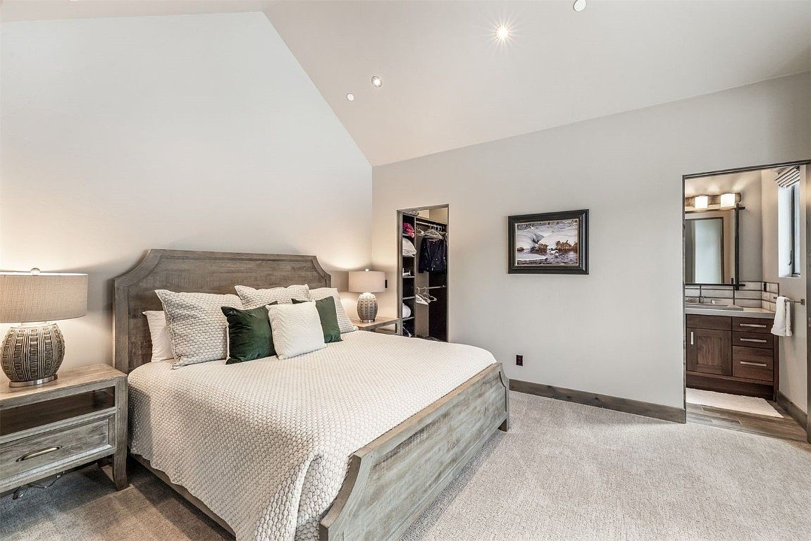 This is a primary bedroom featuring a king-size bed with a light-colored textured comforter and several pillows. The room has a neutral color palette with light gray walls and carpet, complemented by a wooden bed frame and matching nightstands. There is a walk-in closet and a bathroom visible from the bedroom, suggesting an en-suite arrangement.