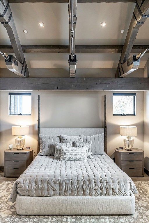 This is a primary bedroom featuring a neutral color palette with a large bed as the focal point. The bed has a light gray upholstered headboard and is covered with a textured quilt and decorative pillows. Two matching nightstands with lamps flank the bed, and small windows are positioned above each nightstand, creating a symmetrical and inviting space.