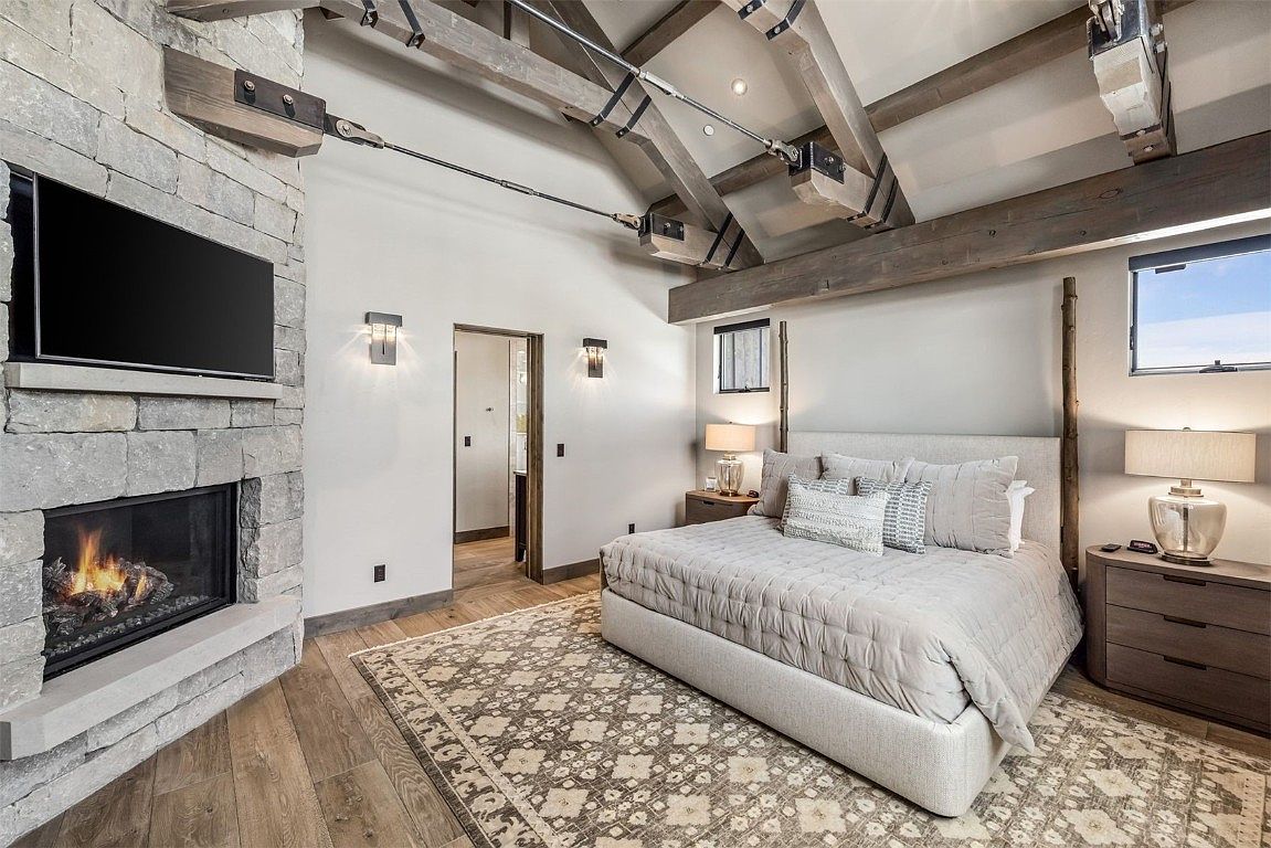 This is a primary bedroom featuring a stone fireplace with a mounted television, a large bed with neutral-toned bedding, and exposed wooden beams on the ceiling. The room has a rustic yet luxurious feel, with hardwood floors and stylish lighting fixtures. The overall impression is one of comfort and sophistication.