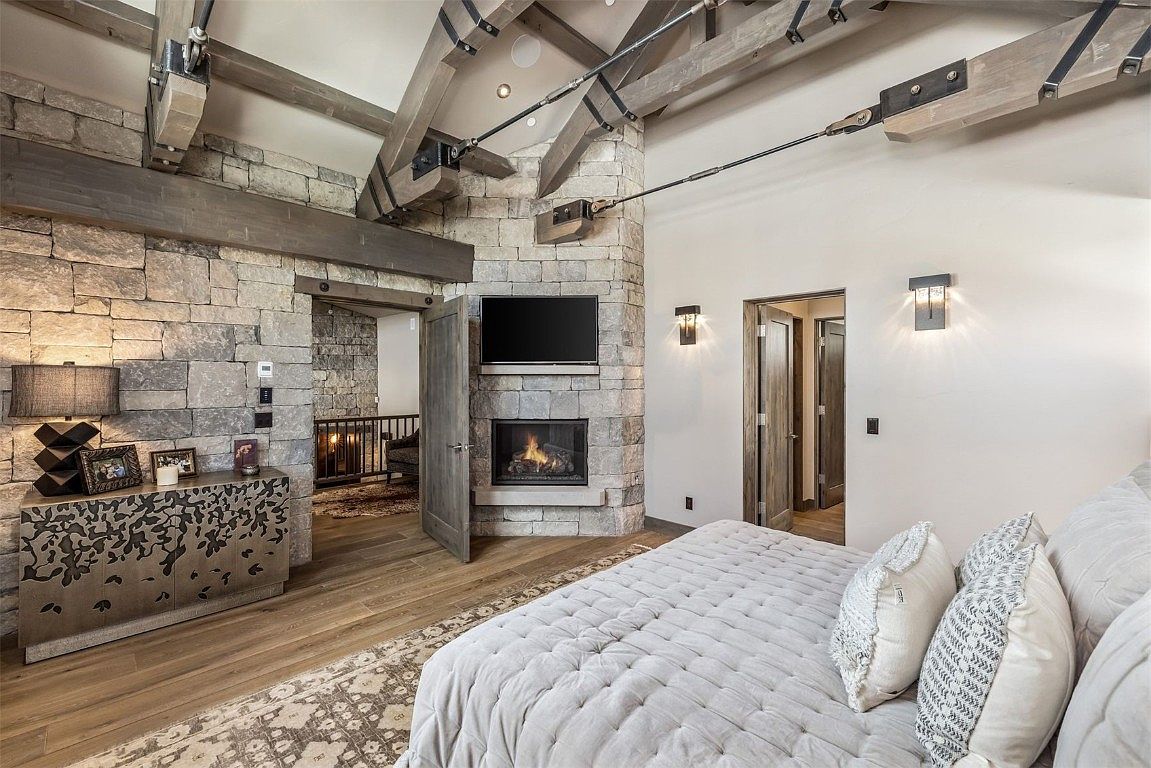 This is a primary bedroom featuring a stone accent wall with a fireplace and mounted TV. The room has exposed wooden beams and a neutral color palette, creating a rustic yet luxurious feel. A large bed with plush bedding is the focal point, and there are doorways leading to other areas of the house.