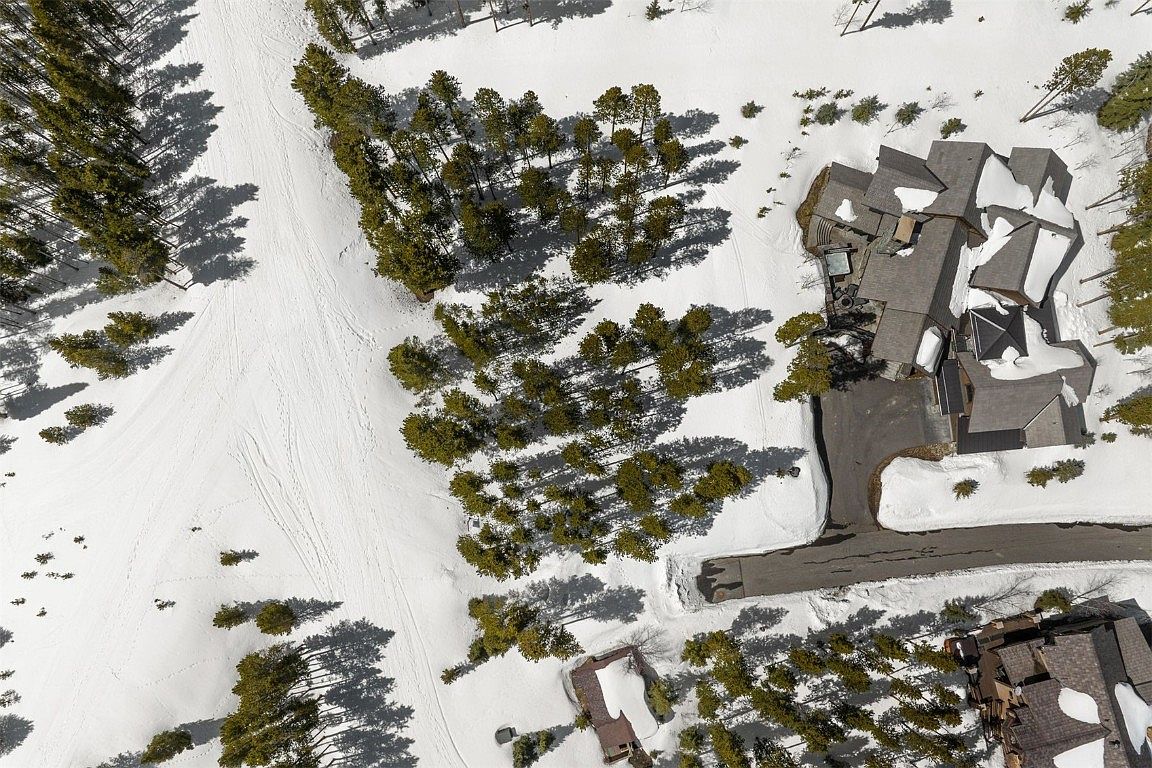 This aerial shot showcases a luxurious mountain home with a complex roofline, nestled among snow-covered terrain and evergreen trees. A winding driveway leads to the property, suggesting privacy and seclusion. The surrounding landscape is a mix of open, snowy areas and dense forest, creating a picturesque winter setting.