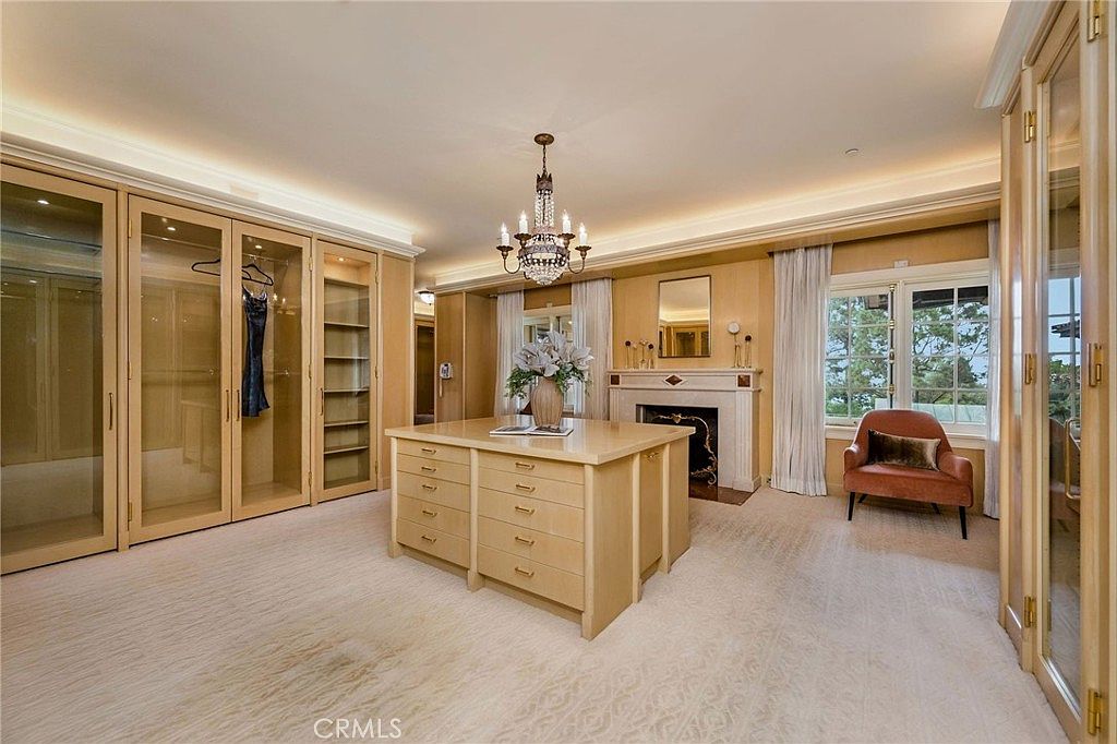 This luxurious walk-in closet features extensive light wood cabinetry with glass-front doors and a central island providing ample storage. A crystal chandelier hangs above the island, while a fireplace with a decorative mantel adds a sophisticated touch to the space. A plush armchair sits near the window, creating a refined and comfortable atmosphere.