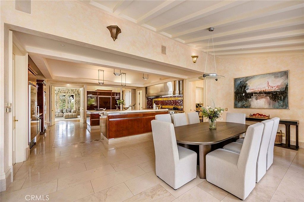 This open-concept space features a large kitchen with a prominent wood-toned island and a dining area set with a dark oval table and light-colored upholstered chairs. The room is characterized by high, beamed ceilings and light-colored stone flooring that flows seamlessly between the cooking and eating zones. A large piece of artwork hangs on the wall near the dining area, while the kitchen is equipped with professional-grade stainless steel appliances and a decorative tile backsplash.