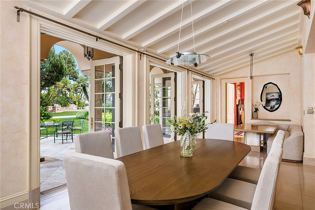 This elegant dining room features a large, oval-shaped wooden table surrounded by upholstered chairs, set beneath a modern, minimalist pendant light. The room is bathed in natural light from expansive glass doors that open directly onto a stone-paved patio and lush garden. A secondary seating area with a built-in bench and a small table is tucked into an alcove, creating a bright and inviting space that seamlessly blends indoor and outdoor living.