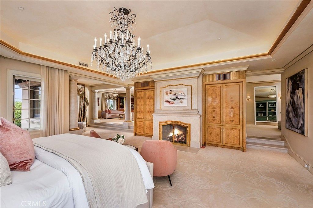 This expansive primary bedroom exudes luxury with its ornate crystal chandelier, tray ceiling, and a central fireplace featuring a decorative mantel. The room includes a raised seating area with columns, plush carpeting, and custom light wood cabinetry flanking the fireplace. Large windows with elegant drapery allow natural light to fill the space, creating a sophisticated and grand atmosphere.
