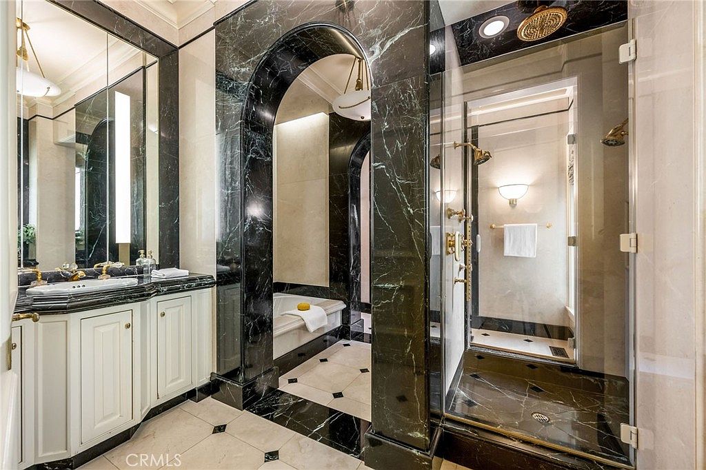 This luxurious primary bathroom features a dramatic arched entryway framed in dark, veined stone that leads to a soaking tub. To the right, a glass-enclosed shower stall is finished with matching dark stone, while a vanity with white cabinetry and a light-toned stone countertop sits to the left. The space is accented by elegant light fixtures and a light-colored tile floor with dark decorative inlays, creating a sophisticated and high-end atmosphere.