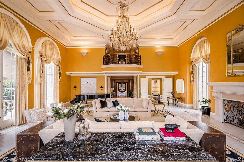 This grand, formal living room features high ceilings with intricate molding, a large crystal chandelier, and vibrant yellow walls. The space is anchored by a plush seating area with a central coffee table, while a dark stone-topped console table in the foreground provides a sophisticated focal point. Large arched windows with elegant drapery flank the room, and a balcony overlooks the main living area from above, creating a sense of opulence and scale.