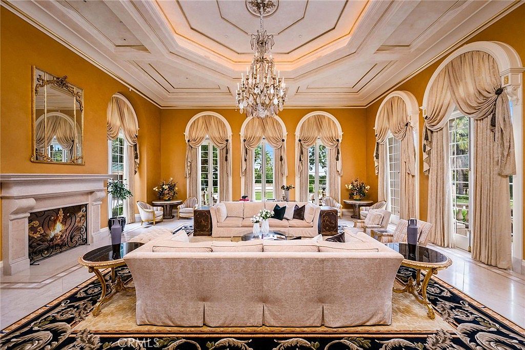 This grand, formal living room features a symmetrical layout with a large, light-colored sofa set centered around a patterned area rug. The space is defined by high, coffered ceilings with an ornate crystal chandelier, tall arched windows draped in elegant fabric, and a prominent stone fireplace on the left. The warm, golden-toned walls and polished flooring create a sophisticated and luxurious atmosphere suitable for formal entertaining.