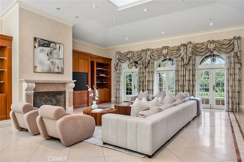 This spacious living room features a neutral color palette with light-toned tile flooring and a large, plush sectional sofa paired with two matching armchairs. A stone-clad fireplace serves as a focal point, complemented by built-in wood cabinetry and shelving. Tall windows draped in ornate, heavy fabric curtains allow natural light to fill the room, creating an elegant and inviting atmosphere.