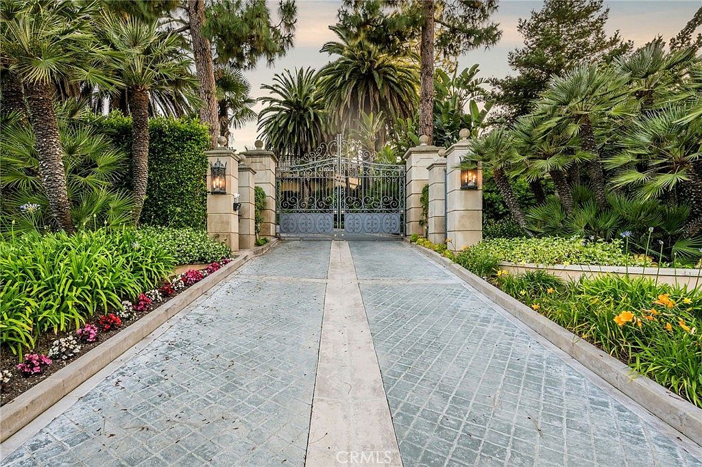This image captures a grand, gated entrance to a private estate, featuring a paved driveway leading up to an ornate, dark metal gate. The entrance is flanked by stone pillars topped with lanterns and surrounded by lush, mature landscaping, including tall palm trees and vibrant flowering plants. The perspective is a low-angle, centered shot that emphasizes the exclusivity and formal curb appeal of the property.