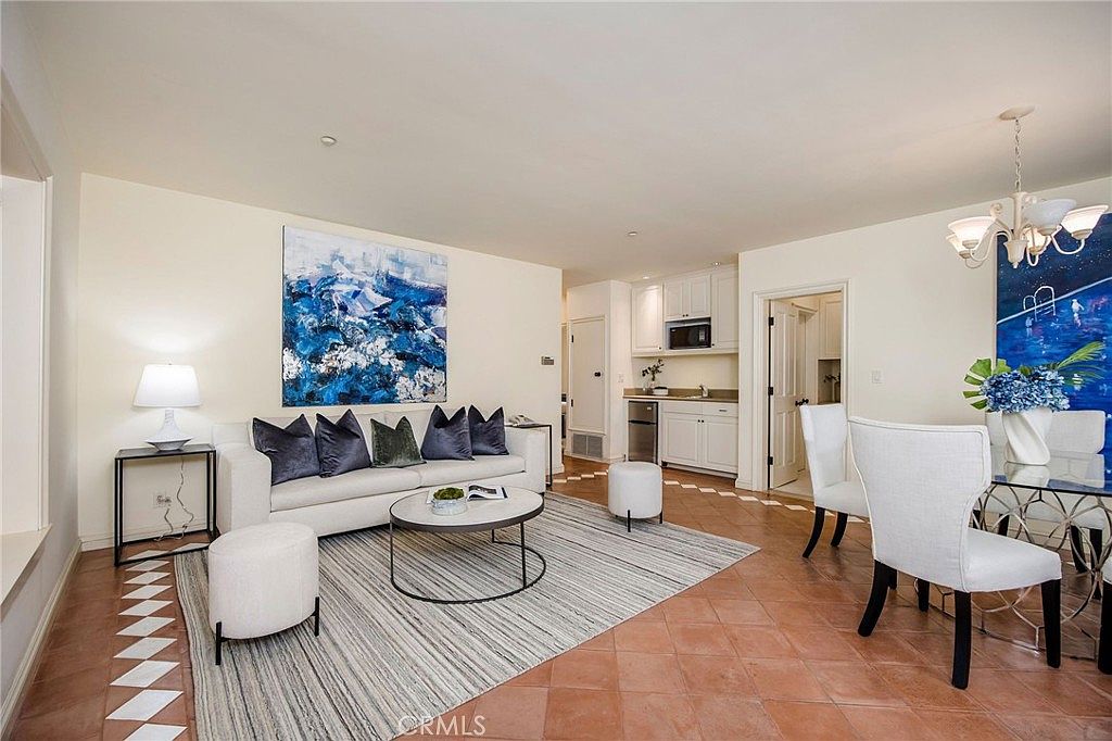 This open-concept living and dining area features warm-toned tile flooring and a neutral color palette accented by bold blue abstract artwork. The living space is anchored by a light-colored sofa with dark throw pillows and a circular coffee table, while the adjacent dining area includes a glass-topped table and white upholstered chairs beneath a decorative chandelier. The space flows seamlessly into a bright kitchen area in the background, creating an inviting and cohesive atmosphere.