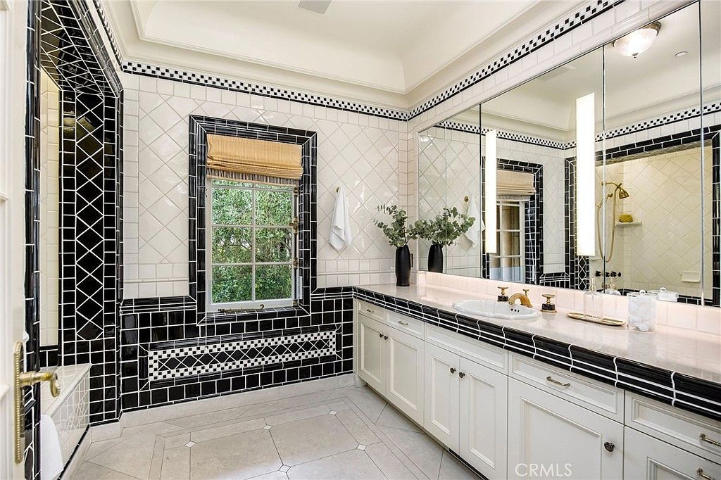 This elegant primary bathroom features a striking black-and-white color scheme with decorative tile work and a classic aesthetic. A long vanity with white cabinetry and a light-toned stone countertop sits beneath a large, multi-paneled mirror, while a window with a woven shade provides natural light. The space is finished with detailed tile borders and high-end fixtures, creating a sophisticated and timeless atmosphere.