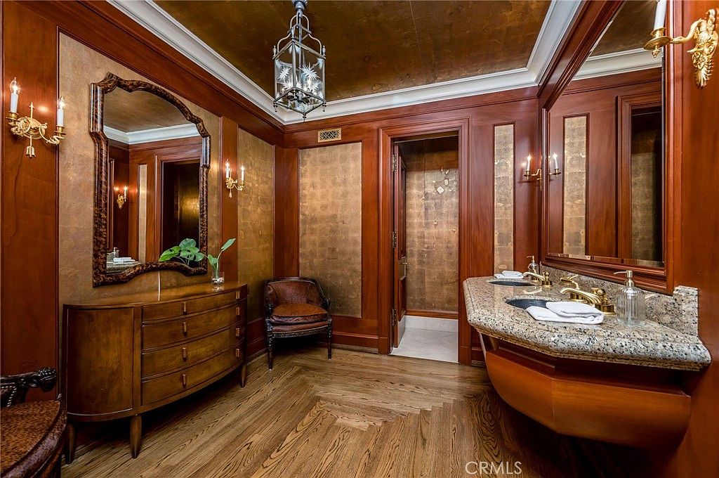 This luxurious primary bathroom features rich wood paneling, gold-toned wall accents, and a warm, traditional aesthetic. The space includes a granite-topped vanity with dual sinks, a decorative mirror, and a classic chandelier, all set against a backdrop of polished wood flooring. A small chair sits in the corner, and a doorway leads to a private water closet, creating an elegant and sophisticated atmosphere.