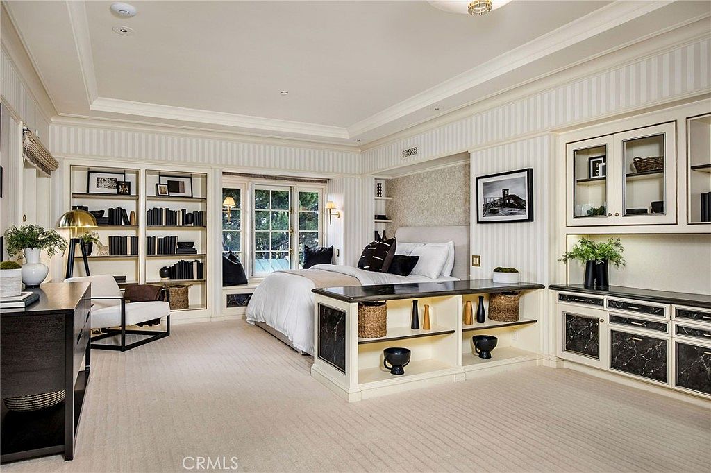 This elegant primary bedroom features a sophisticated design with striped wallpaper, built-in shelving units, and a central bed positioned against a textured accent wall. A long, dark-topped console table sits at the foot of the bed, providing additional storage with woven baskets, while large windows with a window seat offer natural light. The room is finished with neutral carpeting and classic crown molding, creating a refined and tranquil atmosphere.