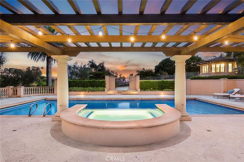 This luxurious outdoor space features a large, rectangular swimming pool paired with a circular, elevated spa in the foreground. A wooden pergola structure with integrated string lighting provides shade over the spa area, supported by classic columns. The scene is set against a sunset sky, with manicured hedges and a stone-walled perimeter creating a private, resort-like atmosphere.