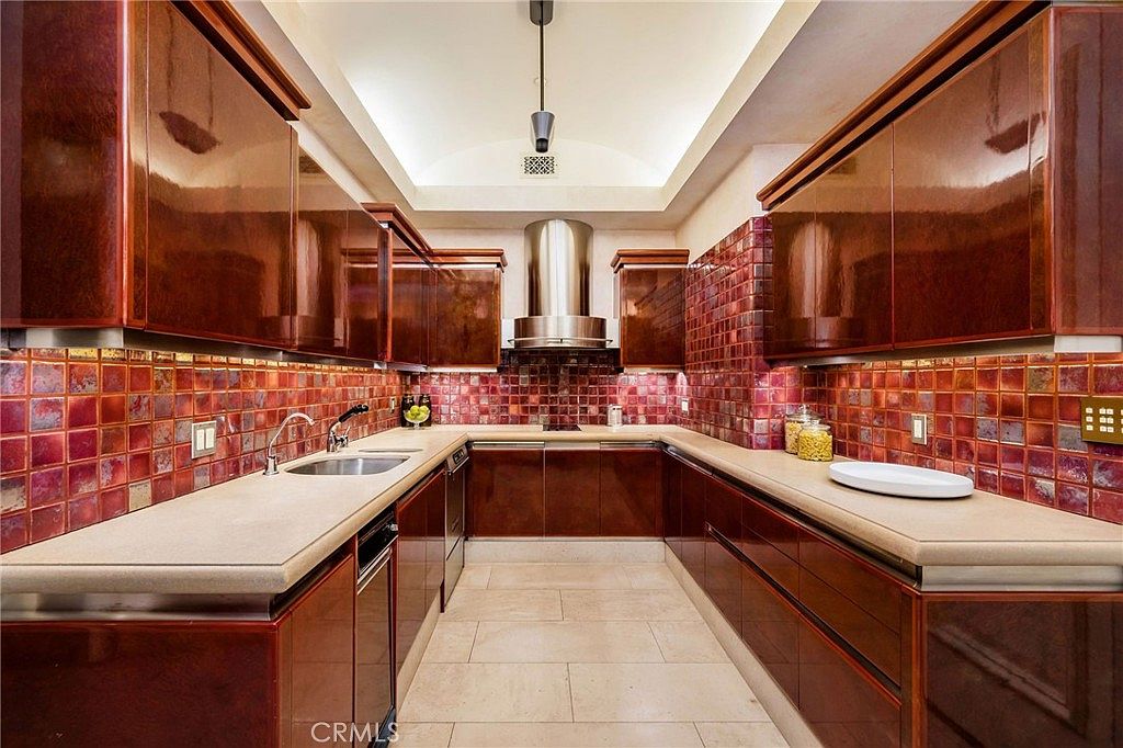 This galley-style kitchen features high-gloss, dark wood-toned cabinetry paired with a distinctive, deep red square-tiled backsplash that covers the walls. The space includes light-colored stone countertops, a stainless steel sink, and a modern range hood centered above the cooktop. The perspective is a straight-on, eye-level shot looking down the length of the kitchen, highlighting the symmetrical layout and the recessed lighting in the cove ceiling.