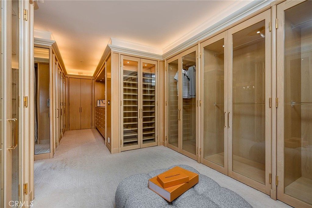 This expansive walk-in closet features custom light wood cabinetry with a mix of open shelving, glass-fronted doors, and integrated drawers. A plush, neutral-toned ottoman sits in the center of the room, providing a focal point atop the wall-to-wall carpeting. The space is illuminated by recessed lighting, creating a warm and organized atmosphere suitable for a luxury primary suite.