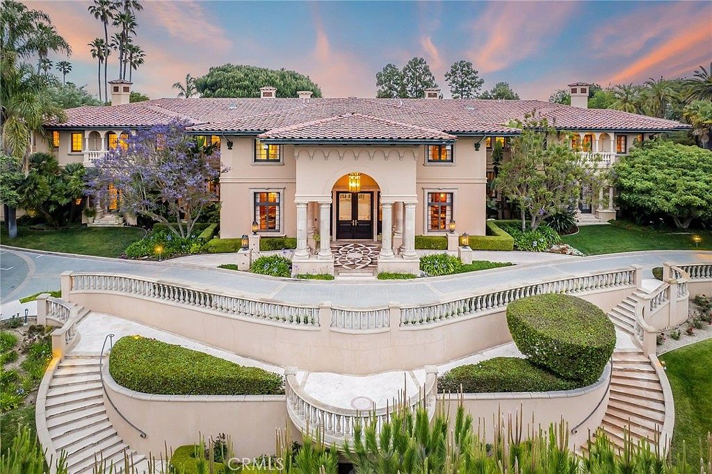 This grand Mediterranean-style estate features a symmetrical facade with a prominent central entryway framed by stone columns and an arched portico. The multi-level exterior is complemented by a sweeping, curved stone staircase that leads down to a landscaped garden area, creating a dramatic and formal arrival experience. The property is surrounded by lush greenery, mature trees, and a paved driveway, all set against a vibrant sunset sky.