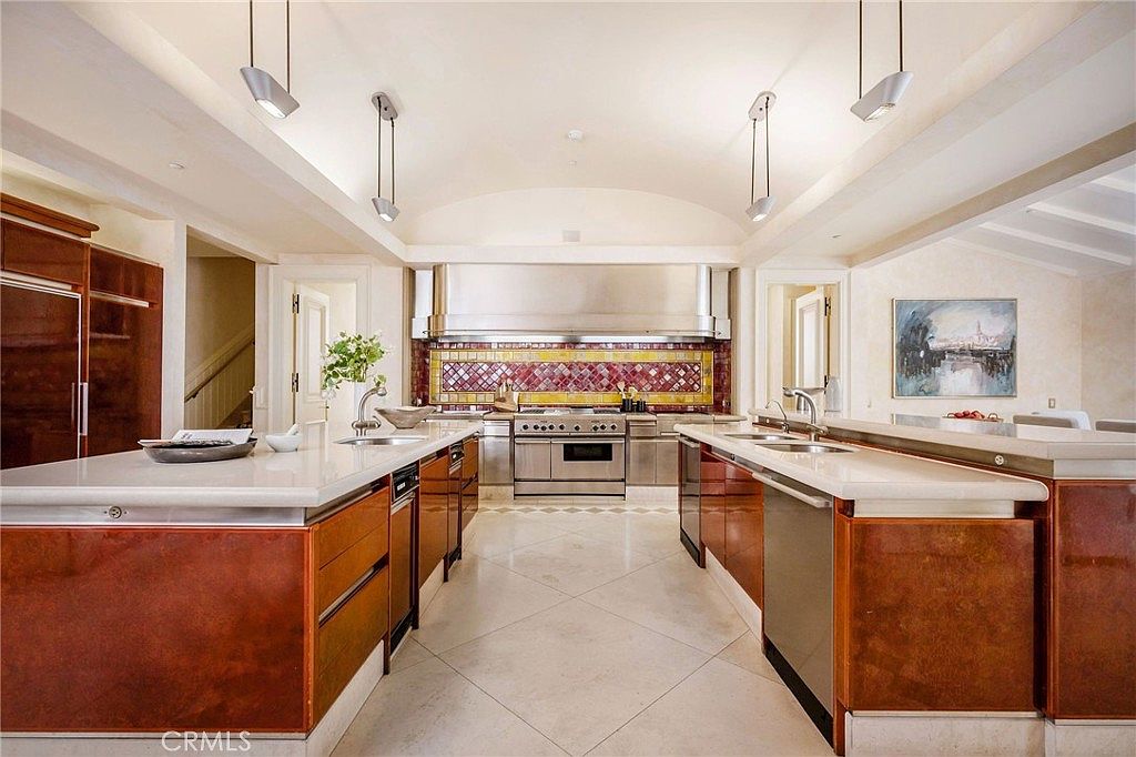 This spacious, symmetrical kitchen features dual islands with light-toned stone countertops and rich wood cabinetry. A professional-grade stainless steel range is centered against a decorative patterned tile backsplash, topped by a large stainless steel hood. The room is illuminated by modern pendant lighting and recessed lights, set against a vaulted ceiling that enhances the open, airy feel of the space.