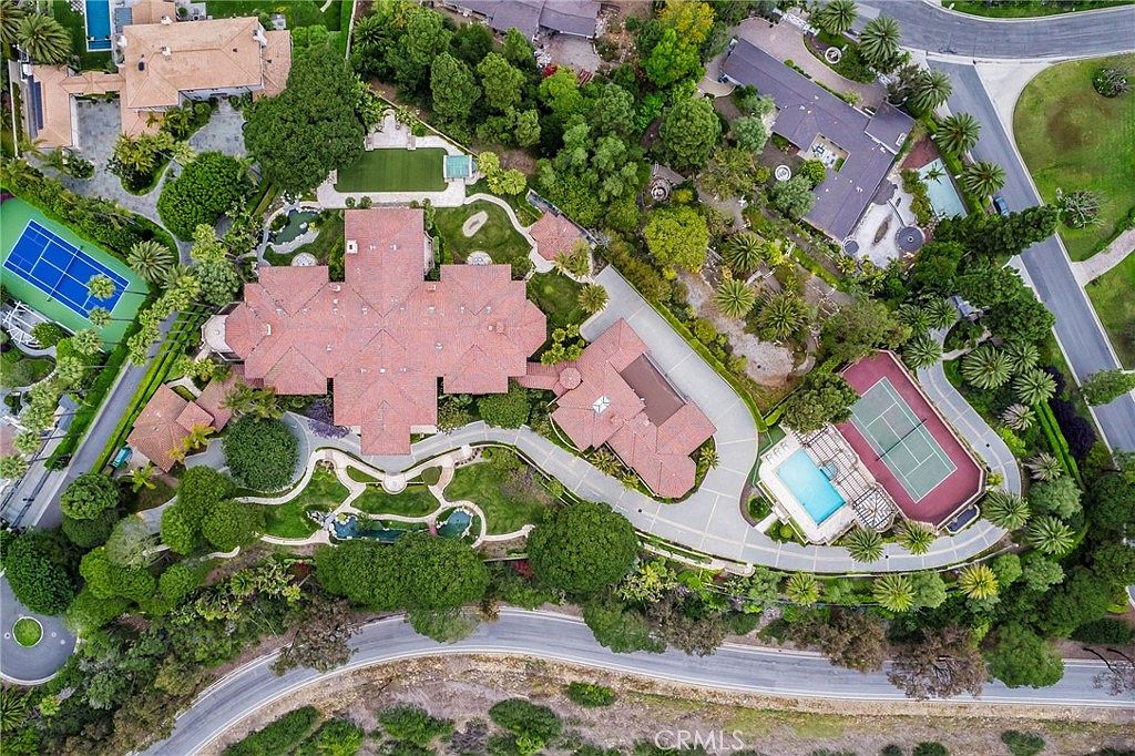 This high-angle aerial view captures a sprawling luxury estate nestled within a lush, tree-lined landscape. The property features a large, multi-winged residence with a terracotta-tiled roof, complemented by a private tennis court, a swimming pool, and extensive manicured grounds. The winding driveway and surrounding greenery emphasize the estate's secluded and expansive nature.