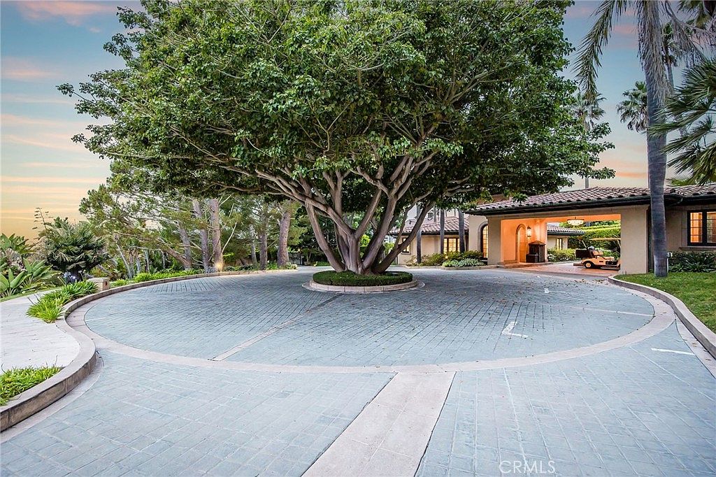 This grand circular driveway features a large, mature tree as its centerpiece, creating a striking focal point for the home's entrance. The paved motor court leads to a covered porte-cochère, providing a sheltered arrival area with warm lighting. The surrounding landscape is lush and well-manicured, contributing to an atmosphere of privacy and luxury.