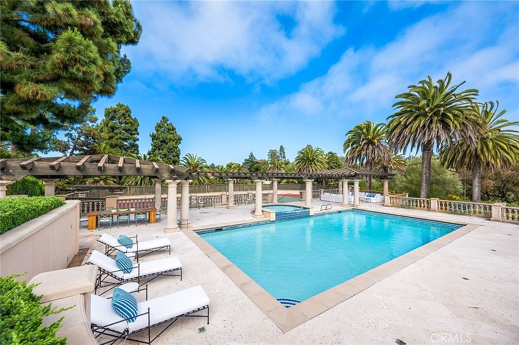 This expansive outdoor living area features a large, rectangular swimming pool with an integrated spa, surrounded by a light-toned stone patio. A wooden pergola structure provides partial shade over a dining area, while several lounge chairs are arranged poolside for relaxation. The scene is set against a backdrop of lush green trees and a clear blue sky, creating a serene and luxurious resort-like atmosphere.