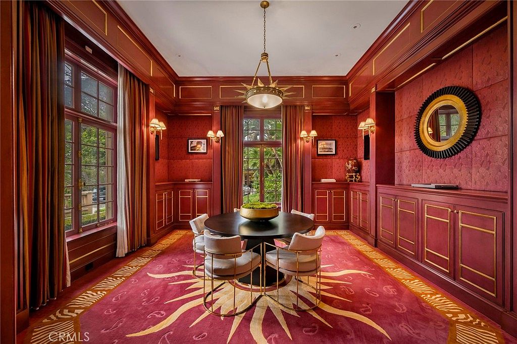 This formal dining room features rich, deep red paneled walls and a striking sunburst-patterned area rug that anchors the space. A central round table is surrounded by four upholstered chairs, illuminated by a decorative gold-toned chandelier. Large windows with floor-length curtains allow natural light to fill the room, creating a sophisticated and dramatic atmosphere.