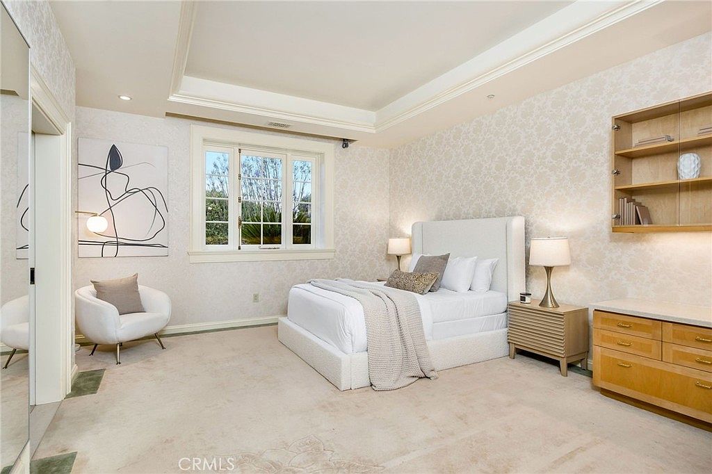 This bright and airy primary bedroom features a neutral color palette with patterned wallpaper and plush carpeting. The room is anchored by a large upholstered bed flanked by matching nightstands and lamps, while a modern white accent chair sits in the corner near a piece of abstract wall art. A built-in light wood shelving unit and matching dresser provide ample storage, and the space is illuminated by a tray ceiling with recessed lighting.