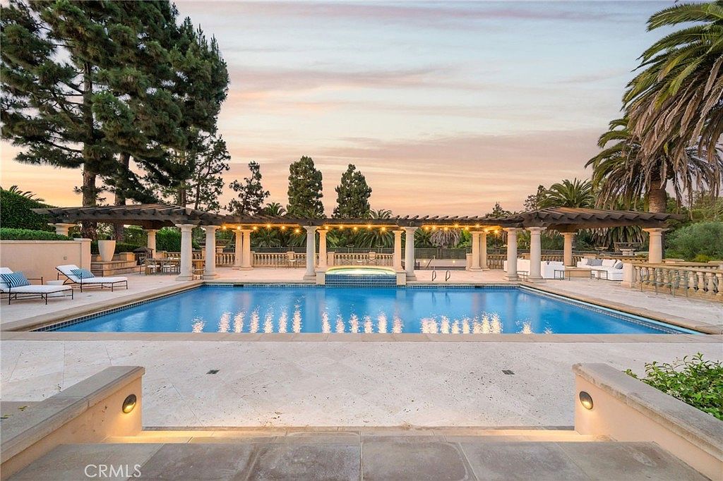 This luxurious outdoor space features a large, rectangular swimming pool with an integrated spa, framed by a classic colonnade pergola. The expansive stone-paved patio is furnished with lounge chairs and outdoor seating, creating an ideal setting for relaxation and entertainment. The scene is captured from a low, centered perspective at dusk, highlighting the warm glow of the pergola lighting against the soft, colorful sunset sky.