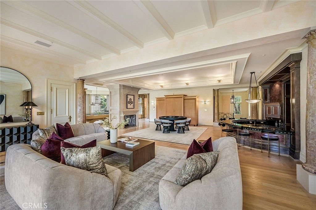This expansive open-concept living area features a sophisticated design with light wood flooring and a coffered ceiling. The space includes a formal seating area with plush sofas, a central game table, and a built-in bar area with dark cabinetry and barstool seating. The room is anchored by a central stone-clad chimney breast, creating a grand and luxurious atmosphere suitable for entertaining.