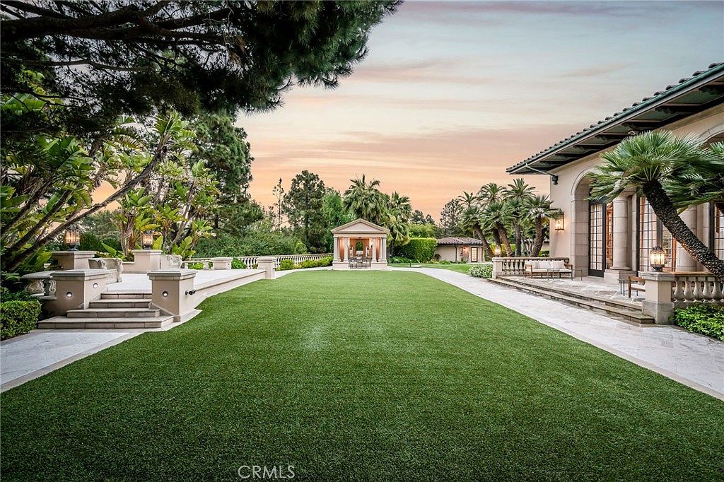 This expansive backyard features a meticulously manicured lawn framed by stone-paved walkways and mature landscaping. A classical-style pavilion stands at the far end of the garden, serving as a focal point, while a covered patio area with outdoor seating extends from the main residence. The scene is captured at dusk, with warm lighting illuminating the architectural details of the home and the garden structures.