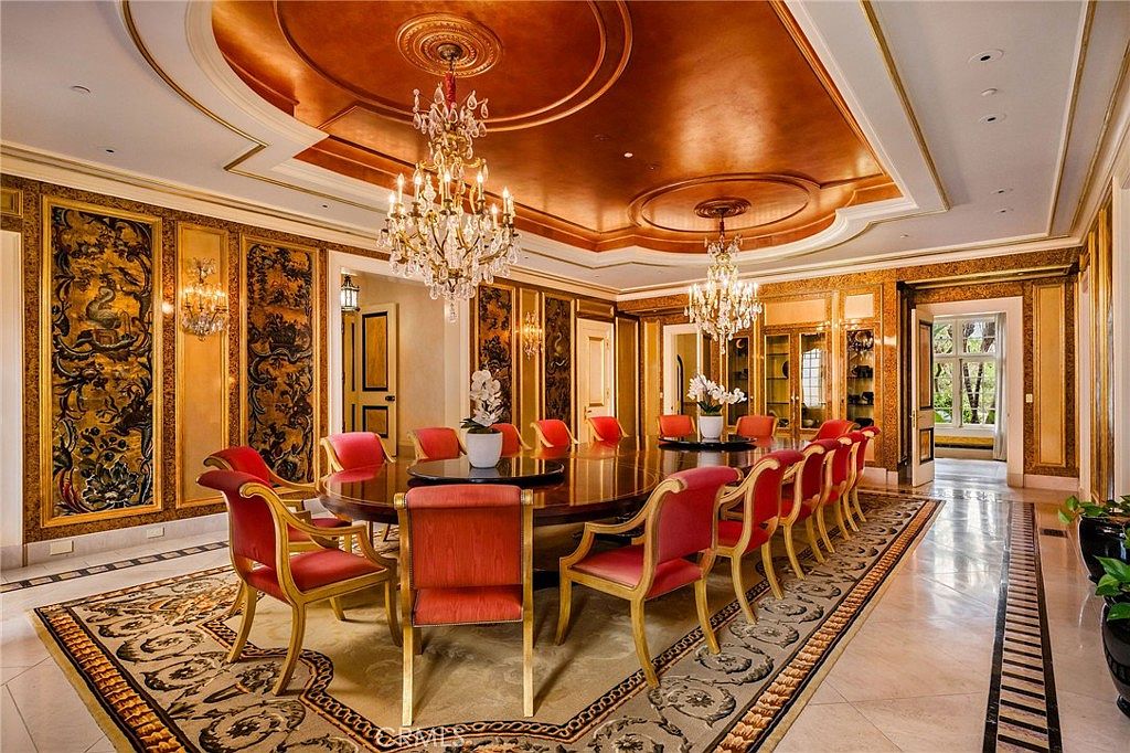 This opulent formal dining room features a large, circular table surrounded by red upholstered chairs, set beneath two ornate crystal chandeliers. The room is defined by its rich, warm color palette, including gold-toned wall panels with intricate dark patterns and a deep copper-colored ceiling. A large, detailed area rug anchors the space, while the polished stone flooring and classic architectural moldings contribute to an atmosphere of grand luxury.