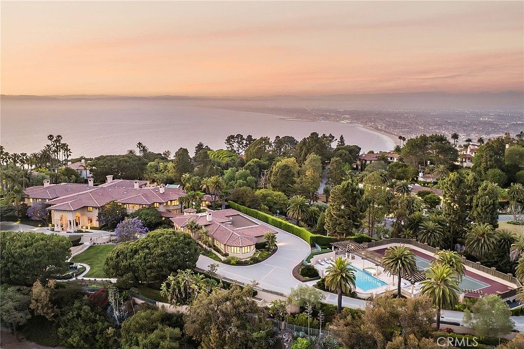 This expansive aerial view captures a luxurious coastal estate set against a breathtaking sunset over the ocean. The property features a sprawling Mediterranean-style residence with multiple wings, a large motor court, and a private swimming pool and tennis court area. Lush, mature landscaping surrounds the home, emphasizing its secluded and prestigious position overlooking the coastline.
