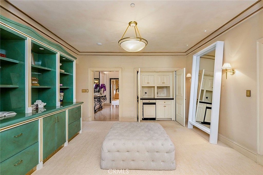 This spacious dressing room or walk-in closet features custom built-in cabinetry in a distinctive teal finish with gold hardware, providing ample storage and display space. A large, tufted ottoman sits in the center of the room, while a tall, white-framed floor mirror leans against the wall, reflecting the adjacent built-in vanity area. The room is illuminated by a classic gold-toned chandelier, creating an elegant and organized atmosphere.